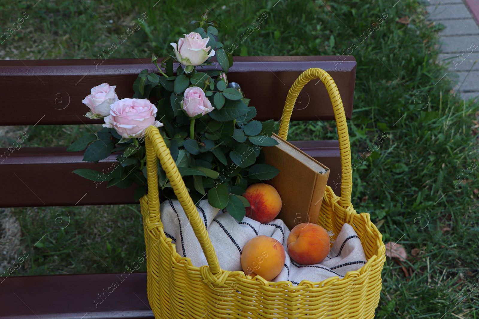 Photo of Yellow wicker bag with roses, book and peaches on bench outdoors