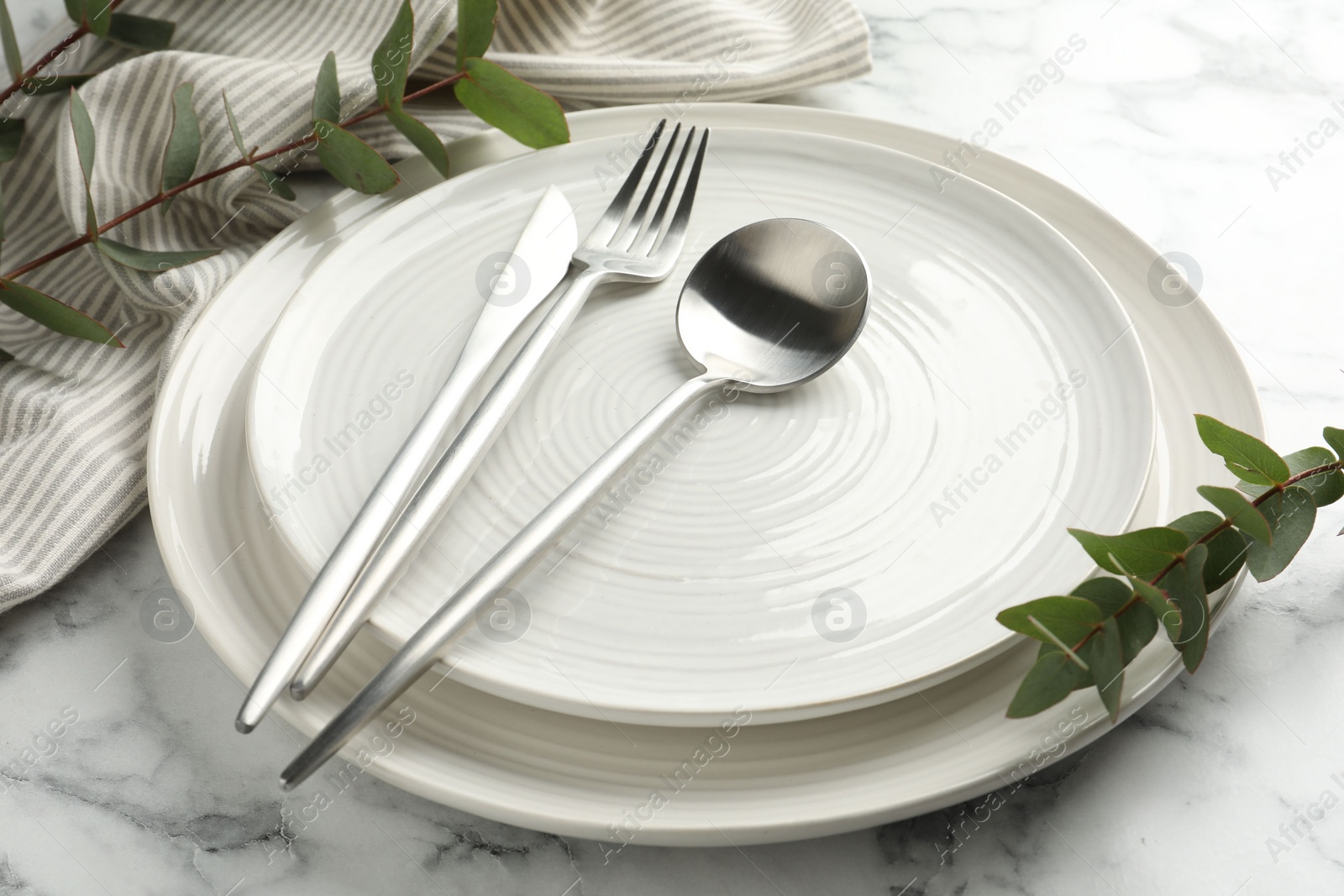 Photo of Stylish setting with cutlery, napkin, eucalyptus branches and plates on white marble table, closeup