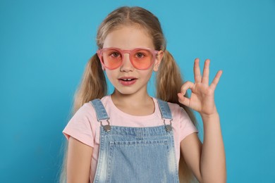 Girl in pink sunglasses showing ok gesture on light blue background