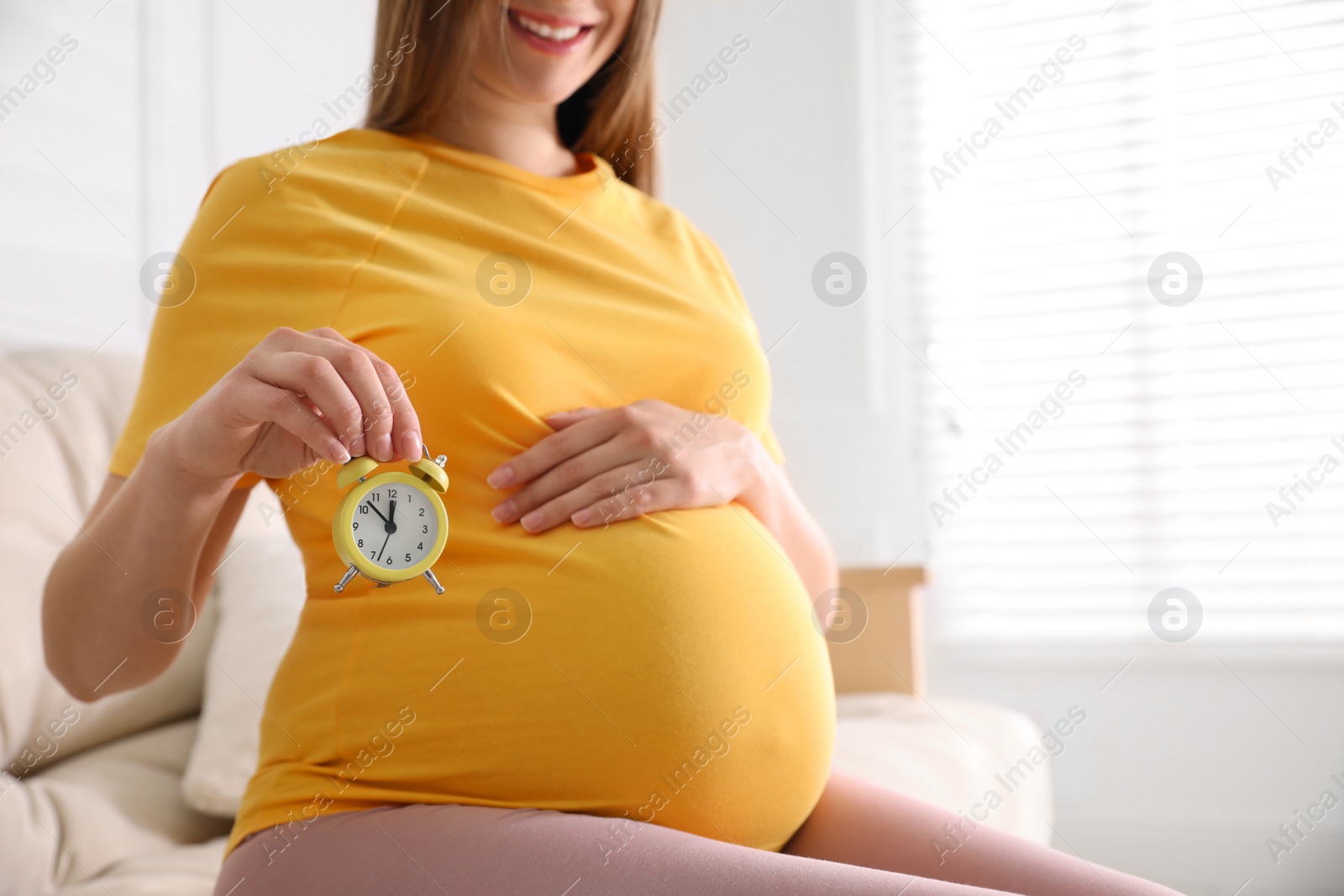 Photo of Young pregnant woman holding alarm clock near her belly at home, closeup. Time to give birth