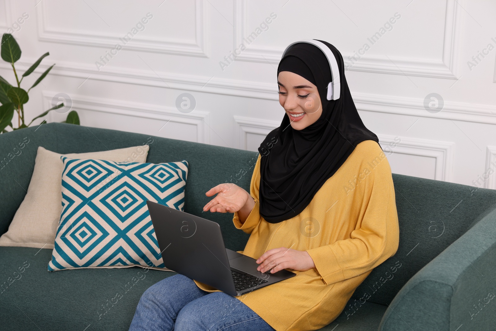Photo of Muslim woman in hijab and headphones using video chat on laptop indoors