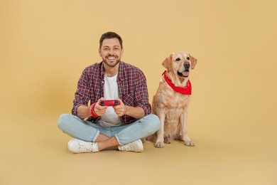 Happy man playing game with controller near his cute Labrador Retriever on beige background