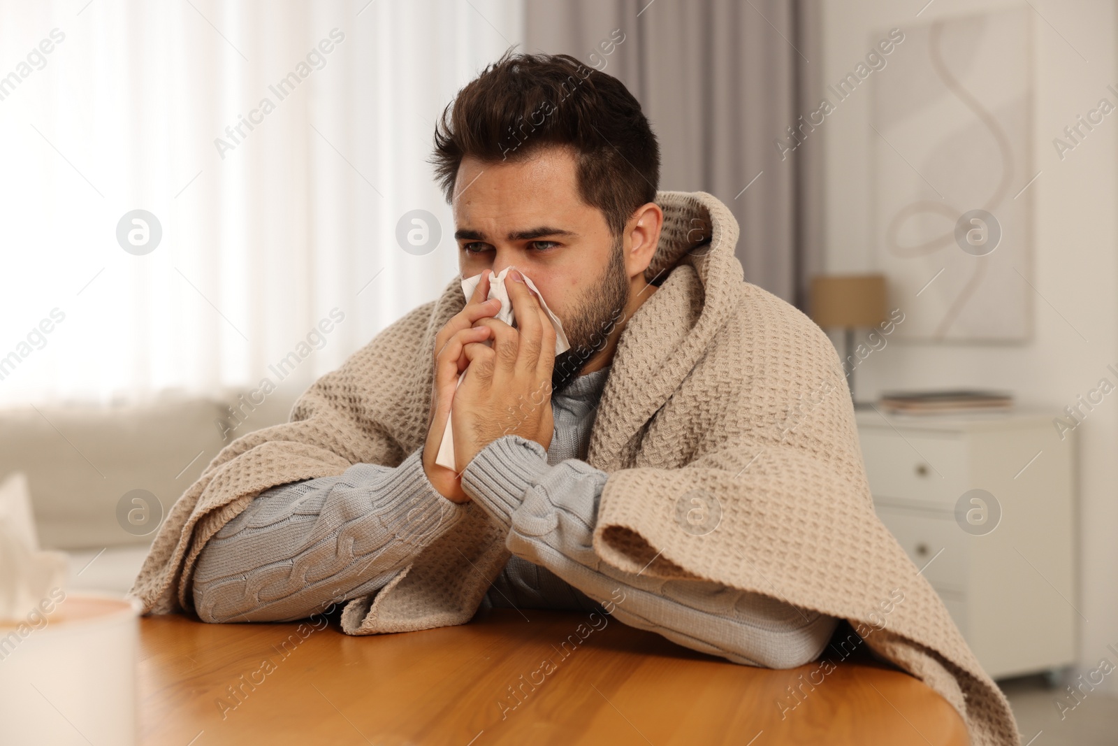 Photo of Sick man wrapped in blanket with tissue blowing nose at wooden table indoors. Cold symptoms