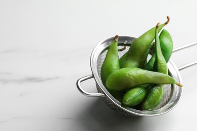 Photo of Sieve with green edamame beans in pods on white marble table. Space for text