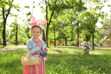 Photo of Little girl with bunny ears holding basket full of Easter eggs outdoors
