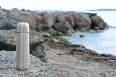 Metallic thermos with hot drink on stone near sea, space for text