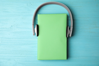 Book with blank cover and headphones on light blue wooden background, flat lay