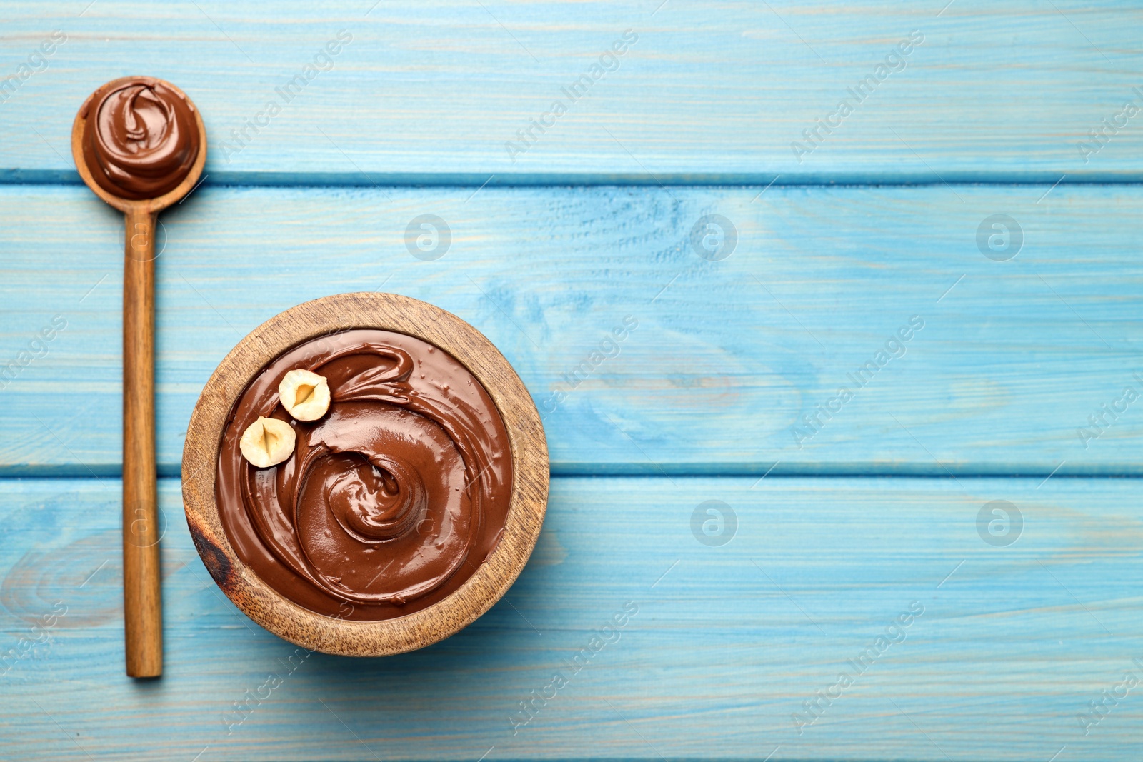 Photo of Spoon and bowl of tasty chocolate hazelnut spread on light blue wooden table, flat lay. Space for text