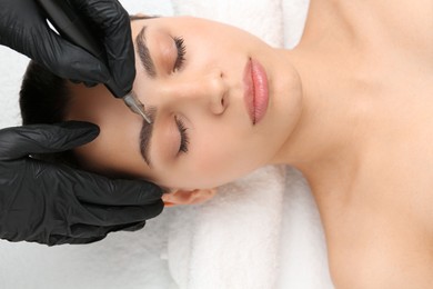 Photo of Beautician making permanent eyebrow makeup to young woman, top view