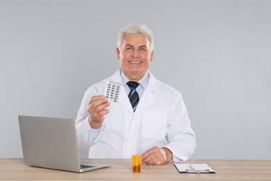 Professional pharmacist with pills and laptop at table against light grey background