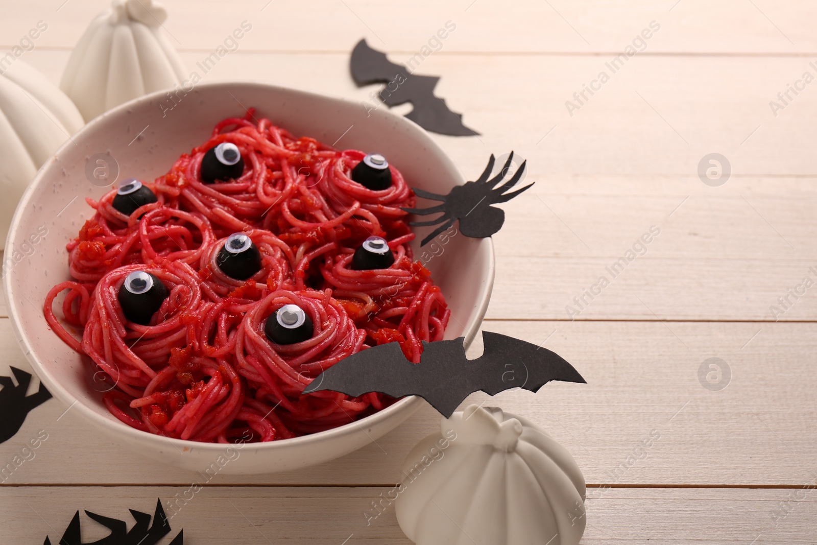 Photo of Red pasta with decorative eyes in bowl and Halloween decorations on white wooden table, closeup. Space for text