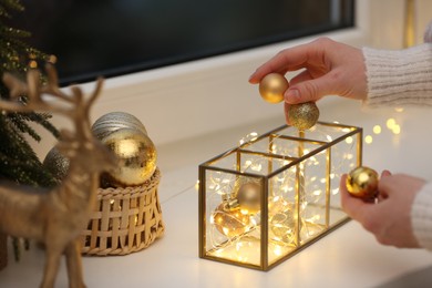 Photo of Christmas atmosphere. Woman putting beautiful baubles into decorative container on window sill indoors, closeup