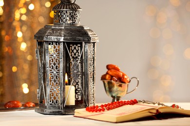 Photo of Arabic lantern, Quran, misbaha and dates on table against blurred lights