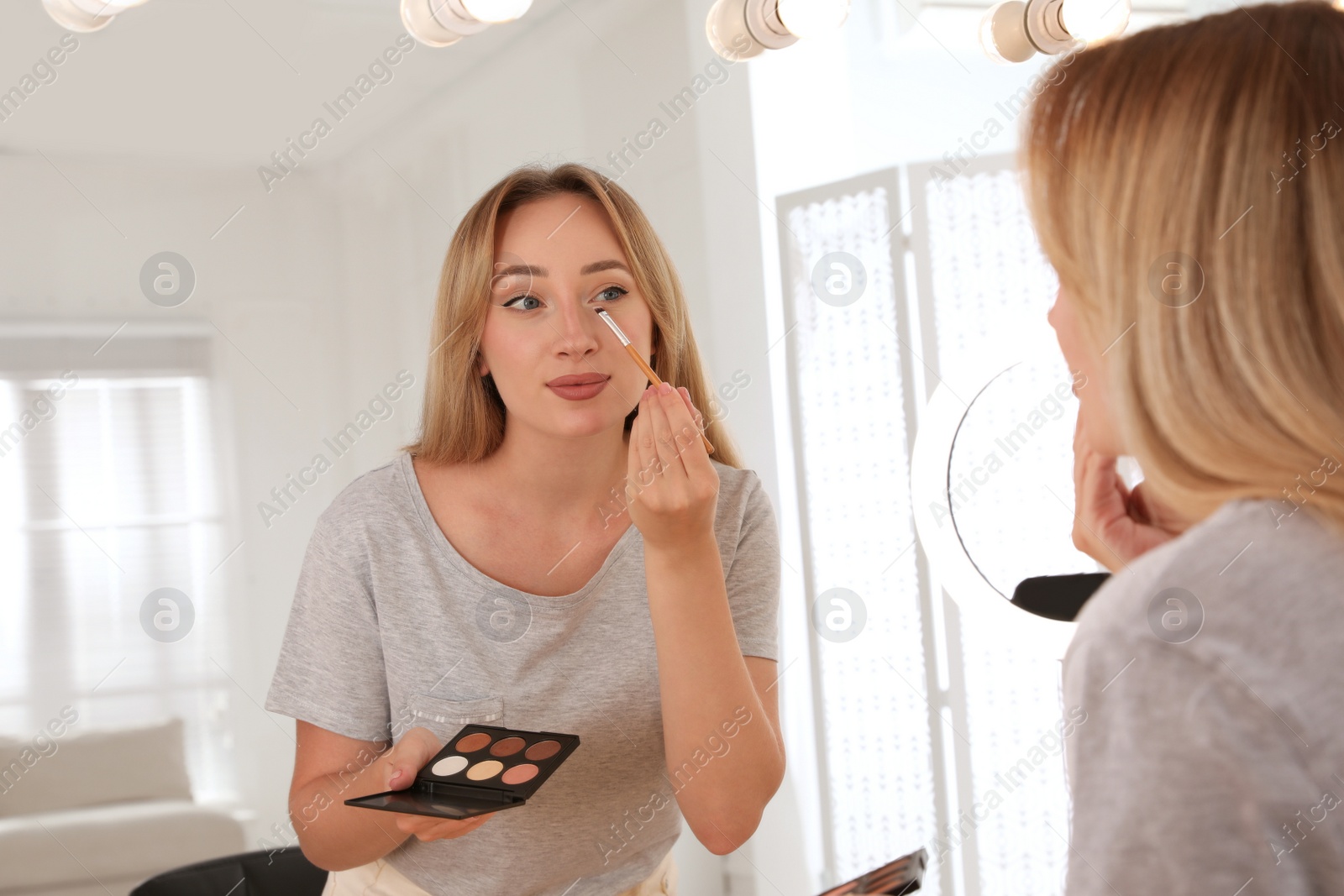 Photo of Beautiful young woman applying eye shadow near mirror and ring lamp indoors