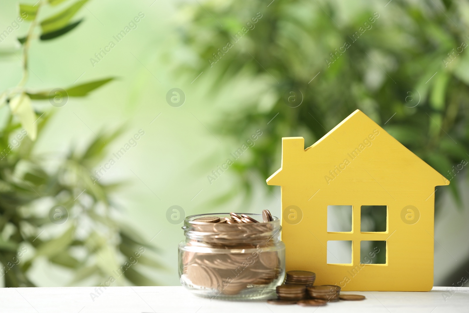 Photo of Model of house and jar with coins on table against blurred green background. Space for text