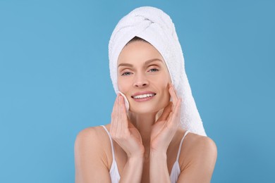 Photo of Beautiful woman in terry towel removing makeup with cotton pad on light blue background