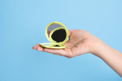 Woman holding stylish cosmetic pocket mirror on light blue background, closeup