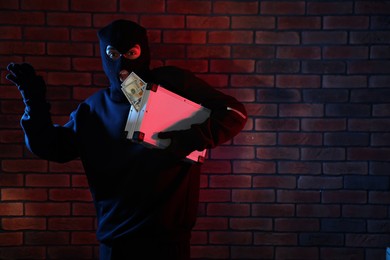 Emotional thief in balaclava with briefcase of money in color lights against red brick wall. Space for text