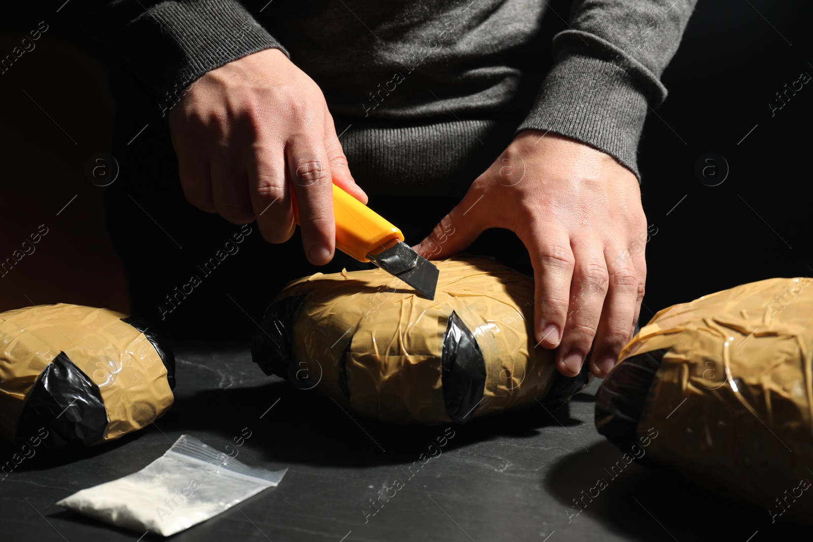 Photo of Smuggling and drug trafficking. Man opening package of narcotics with box cutter at dark table, closeup