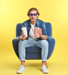 Photo of Emotional man with 3D glasses, popcorn and beverage sitting in armchair during cinema show on color background