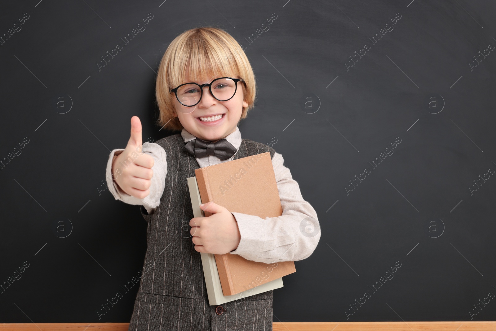 Photo of Happy little school child with notebooks showing thumbs up near chalkboard. Space for text