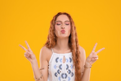 Photo of Beautiful young hippie woman showing V-sign and sending air kiss on orange background