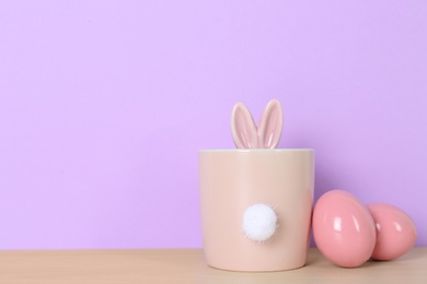 Dyed eggs near cute pot with Easter bunny ears and tail on table against color background, space for text