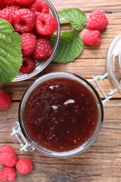 Photo of Jar of delicious raspberry jam, fresh berries and green leaves on wooden table, flat lay