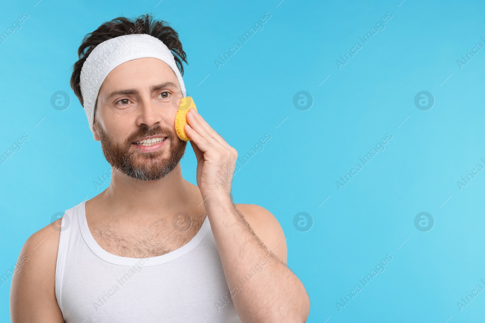 Photo of Man with headband washing his face using sponge on light blue background, space for text