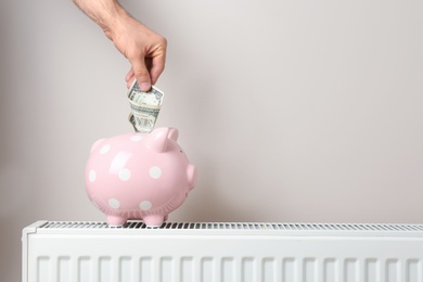 Photo of Man inserting money into piggy bank on heating radiator against color background. Space for text