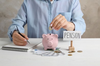 Photo of Pension savings. Woman writing in notebook and putting coin into piggy bank at white wooden table, closeup