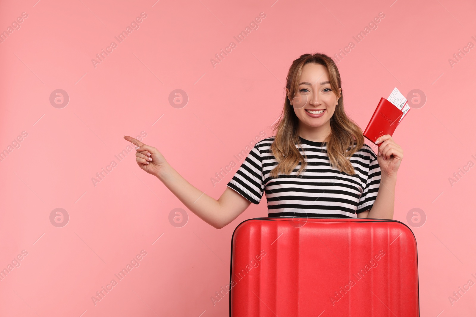 Photo of Happy young woman with passport, ticket and suitcase pointing at something on pink background, space for text