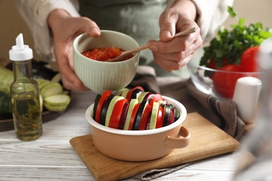 Photo of Cooking delicious ratatouille. Woman dressing fresh vegetables in bowl at white wooden table, closeup