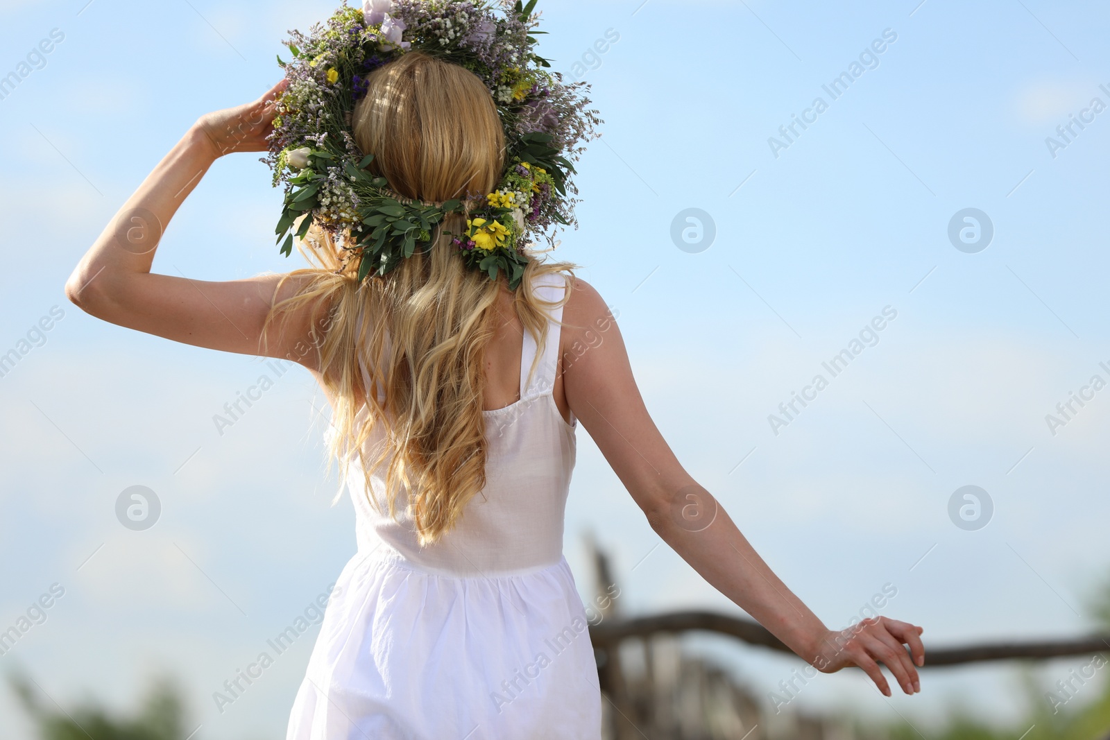 Photo of Young woman wearing wreath made of beautiful flowers outdoors on sunny day, back view