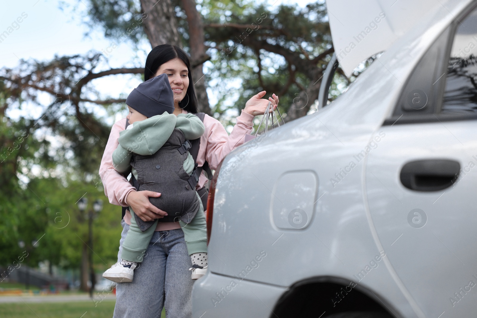 Photo of Mother holding her child in sling (baby carrier) while putting shopping bags into car trunk outdoors