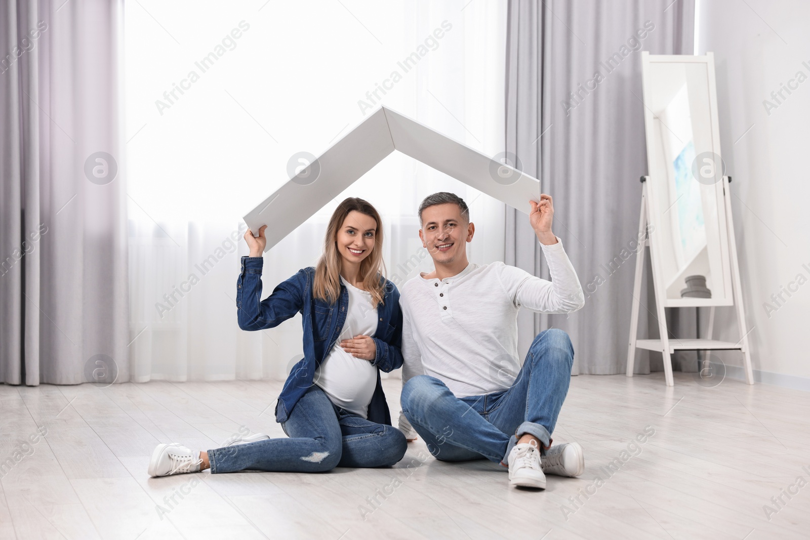 Photo of Young family housing concept. Pregnant woman with her husband sitting under cardboard roof on floor at home