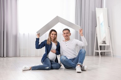 Photo of Young family housing concept. Pregnant woman with her husband sitting under cardboard roof on floor at home