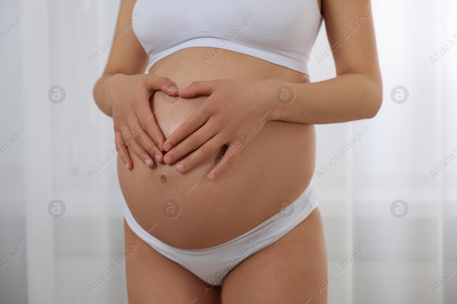 Photo of Pregnant woman in stylish comfortable underwear making heart with hands on her belly indoors, closeup