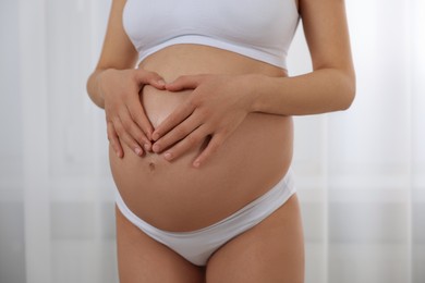 Pregnant woman in stylish comfortable underwear making heart with hands on her belly indoors, closeup