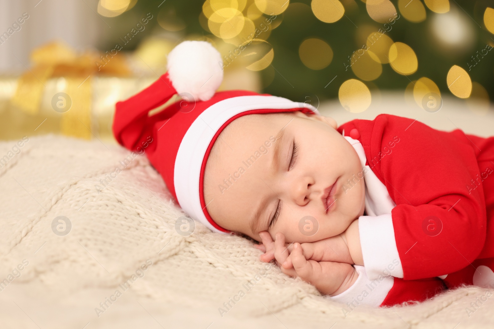 Photo of Cute baby in Christmas costume sleeping on knitted blanket against blurred festive lights. Winter holiday