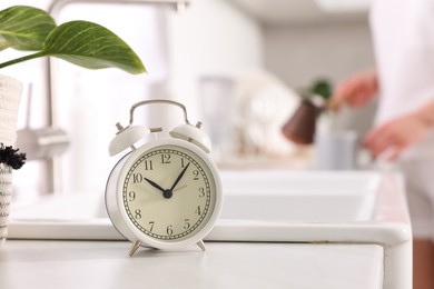 Photo of Alarm clock on white countertop. Woman pouring coffee from jezve into cup in kitchen, selective focus