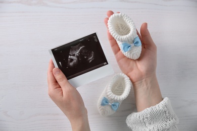 Photo of Woman holding ultrasound photo of baby and knitted boots over table, top view