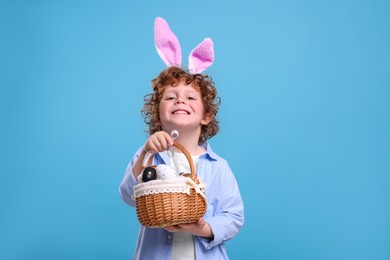 Photo of Portrait of happy boy in cute bunny ears headband holding wicker basket with Easter eggs on light blue background