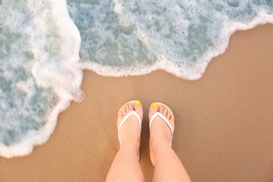 Photo of Top view of woman with white flip flops on sand near sea, space for text. Beach accessories