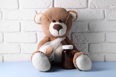 Photo of Toy bear with bandage and bottle of pills on light blue table near white brick wall