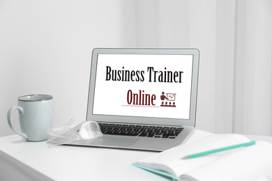 Image of Modern laptop with text BUSINESS TRAINER ONLINE on desk indoors