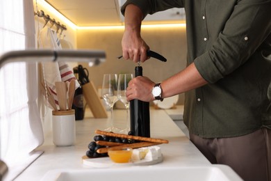 Photo of Romantic dinner. Man opening wine bottle with corkscrew at countertop in kitchen, closeup