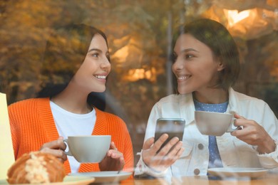 Photo of Special Promotion. Happy young women with cups of drinks using smartphone at table in cafe, view from outdoors