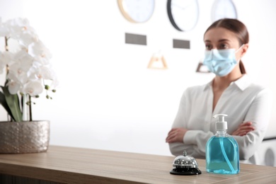 Photo of Receptionist at countertop in hotel, focus on dispenser bottle with antiseptic gel and service bell. Space for text