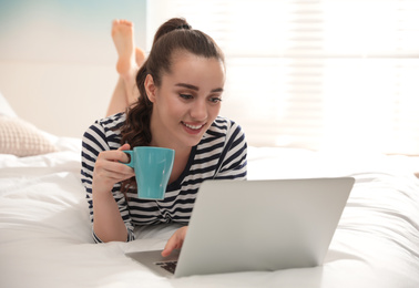 Photo of Young woman with cup of coffee and laptop on bed at home